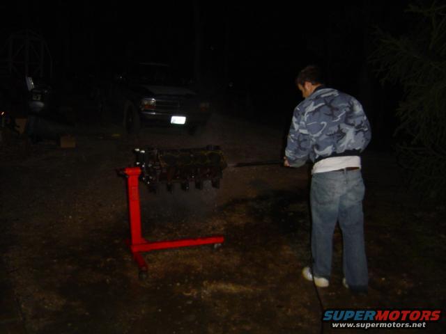 Dark SUV with a white license plate, viewed from the front at night. An engine block is on a red stand in the foreground