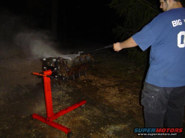 Engine block on a red stand, being sprayed with water by a person in a blue shirt