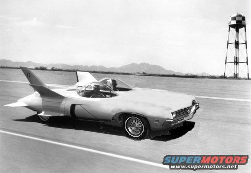 Experimental jet car with two cockpits on a paved surface, water tower in background