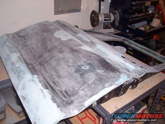 Car door being worked on, covered in primer and body filler. Grinding wheel and tool chest in background