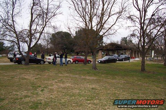 Several cars parked in a park with a picnic shelter and trees