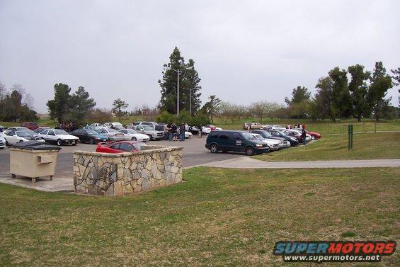 Parking lot with many cars and a green van, stone wall, and dumpster