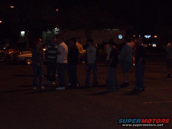 Group of people gathered around cars in a parking lot at night. A motorcycle is visible on the left