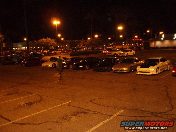 Several cars parked in a lot at night, including a white car with a black hood