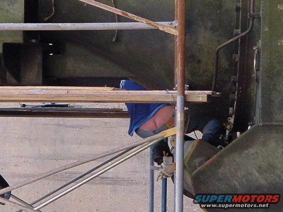 Person working on vehicle under scaffolding. Supermotors.net logo visible