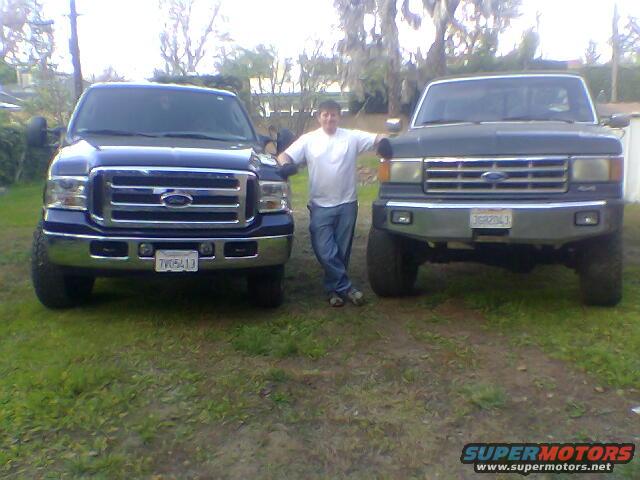 Two Ford trucks, one dark blue and one dark gray, parked on grass. A person stands between them. The blue truck is newer, the gray truck is older
