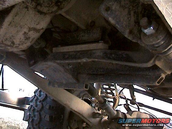 Underbody view of a vehicle showing suspension components and large tires. Watermark reads SUPERMOTORS.NET