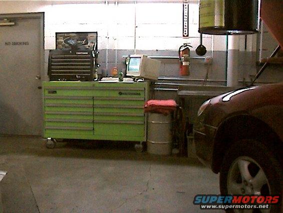 Car fender and wheel in an auto repair shop, with tool chests and a computer visible