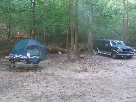 Dark blue SUV parked in a wooded campsite next to a tent and picnic table. The SUV appears to be a 1995 Ford C-150 Centurion Conversion