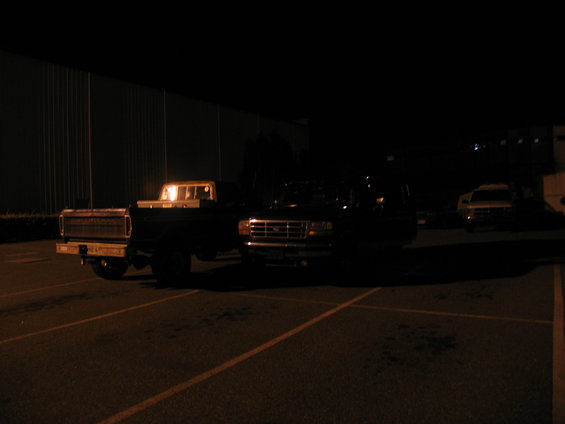 Two dark-colored Ford trucks parked at night. The truck on the left is older, with a chrome bumper and grille. The truck on the right is newer, with a prominent grille and headlights
