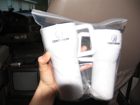 Two white travel mugs with Centurion logo inside a plastic bag, held by a hand. Interior of a vehicle is visible in the background