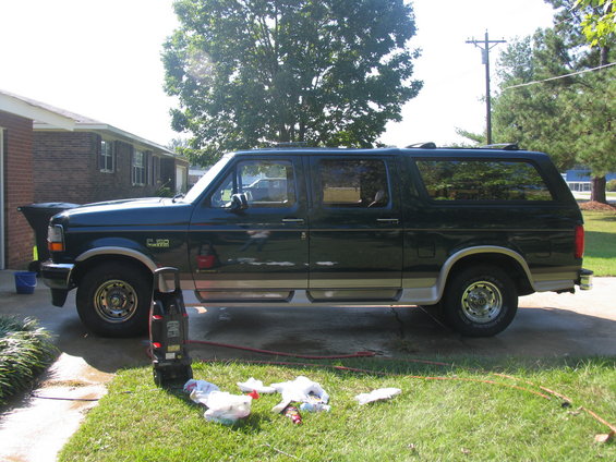 Dark green SUV with tan trim, side view, parked on driveway with pressure washer and cleaning supplies on lawn