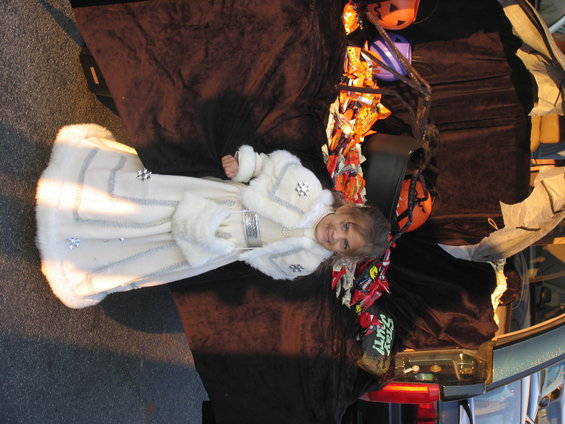 Girl in a white costume with fur trim sits in a car trunk decorated for Halloween with candy and pumpkins