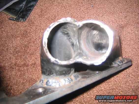 close-up of a welded exhaust manifold with two openings, resting on a brown carpeted surface