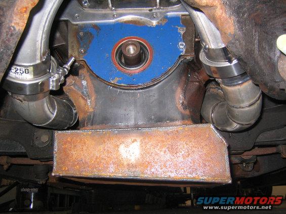 Engine block with blue plate, red seal, and stainless steel pipes. Rusty metal shield below