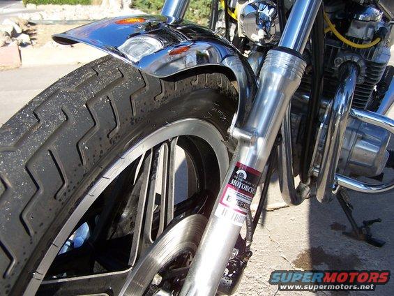 Close-up of a motorcycle's front tire, chrome fender, and suspension forks. A sticker on the fork reads 'MOTORCYCLE'