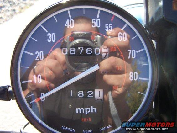 Motorcycle speedometer with odometer reading 1821, reflecting a person holding a camera