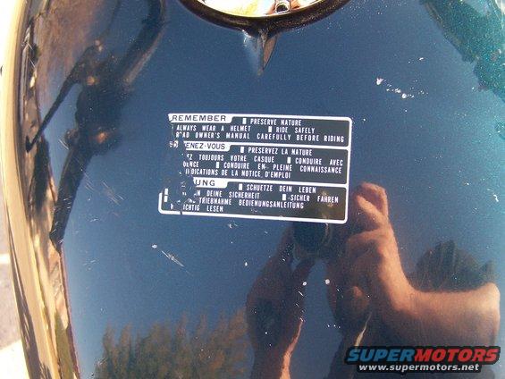Close-up of a sticker on a dark green motorcycle fuel tank with safety instructions in English, French, and German