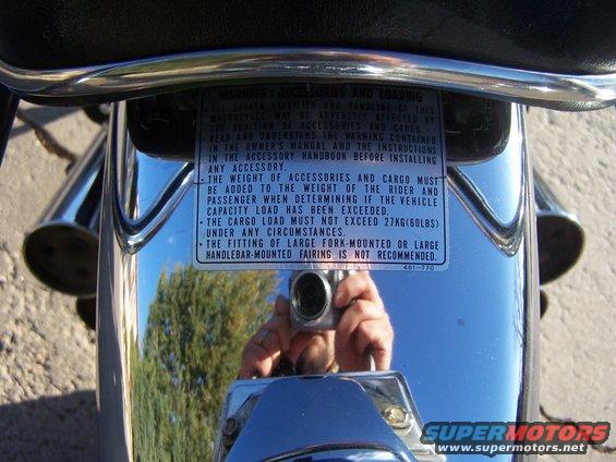 Motorcycle rear fender with a warning label about accessories and loading, and a reflection of the photographer