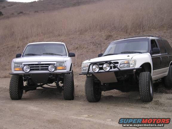 Two modified Ford Explorer SUVs parked on a dirt road, one white, one white and black