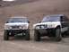 1985 Ford Bronco II - Two modified Ford Explorer SUVs with off-road bumpers and large tires are parked on a dirt road.