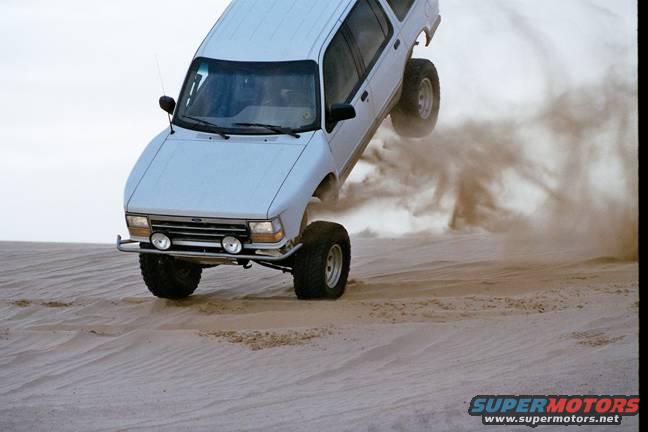 White SUV with lifted suspension and large tires airborne over sand, kicking up dust. Front grille and bumper visible