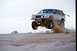 1985 Ford Bronco II - A modified SUV soars through the air above sandy terrain, creating a cloud of dust.