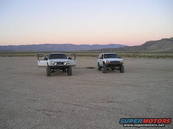 Two white SUVs parked in a dry lakebed at dusk. The SUV on the left has its doors open