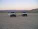 1985 Ford Bronco II - Two SUVs are parked in a vast, dry, cracked earth landscape with mountains in the background at sunset.