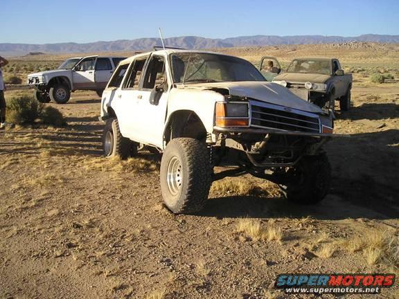 White SUV with lifted suspension and off-road tires parked in a desert landscape with two trucks behind it