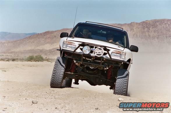 White SUV with off-road tires and a brush guard driving on a dirt road, kicking up dust