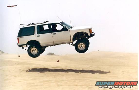 White SUV airborne over sand dunes, driver visible inside