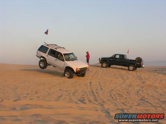 White SUV airborne over sand dune, black pickup truck on dune crest