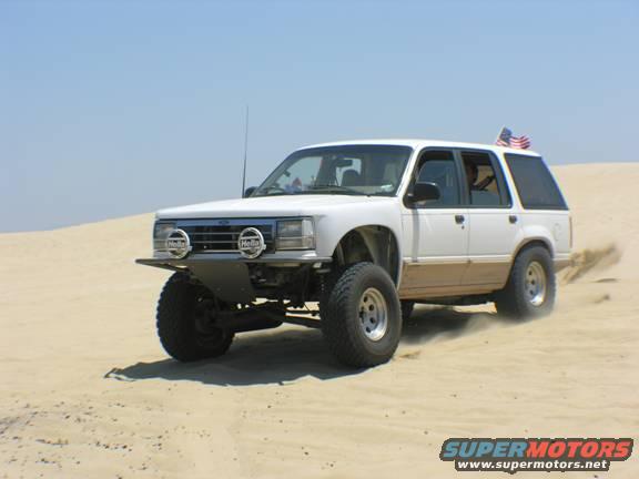 White SUV with lifted suspension and off-road tires driving on sand dunes, kicking up dust