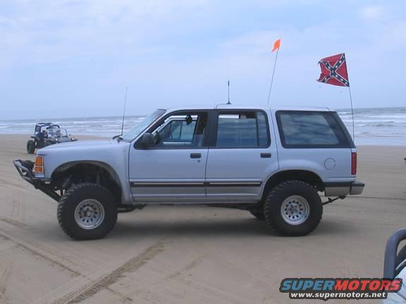 Silver SUV with off-road tires on a beach, another vehicle in background. Flags on poles