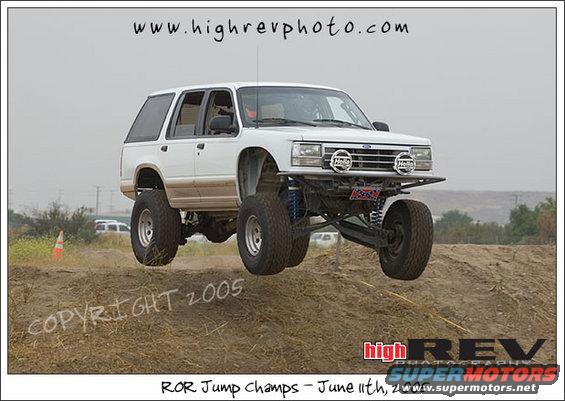White SUV with lifted suspension mid-air over a dirt mound. It has large off-road tires and auxiliary lights on the front bumper