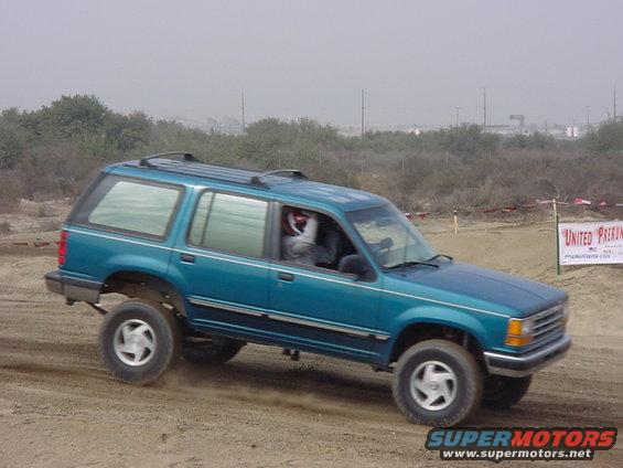 Teal SUV with roof rack driving on dirt, driver wearing red