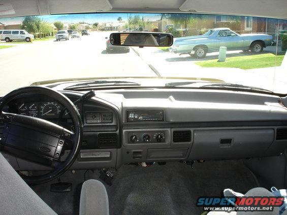 Dashboard of a light blue 1992 Ford F-150 truck, viewed from the passenger seat. The steering wheel and dashboard are visible, along with the street and houses outside