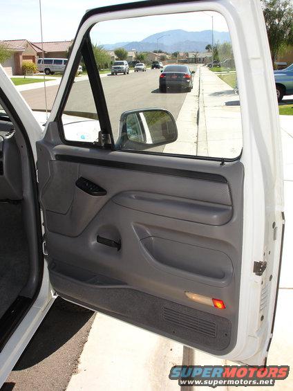 Interior door panel and side mirror of a 1996 Ford Bronco, showing the gray door trim, window controls, and a reflection of the street in the mirror