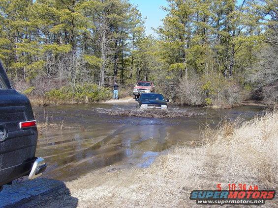 Trucks driving through mud and water on a wooded trail. One truck is in the foreground, partially visible