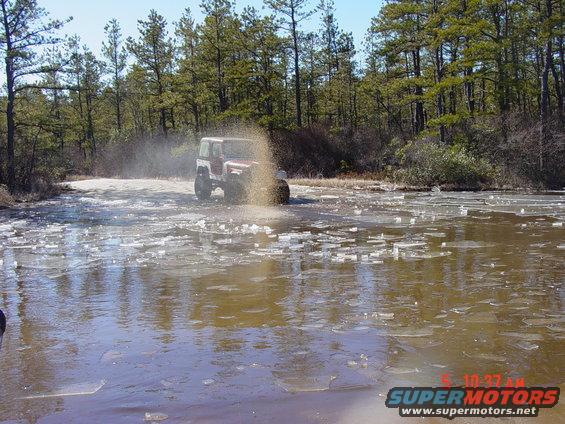 Red and white Jeep Wrangler driving through shallow water with ice chunks, kicking up spray