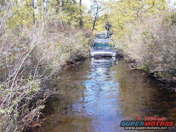 1995 Dodge 1500 and another truck driving through water in a wooded area