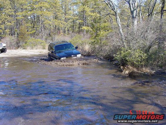 Black SUV stuck in a muddy water crossing. Another vehicle is visible on the bank