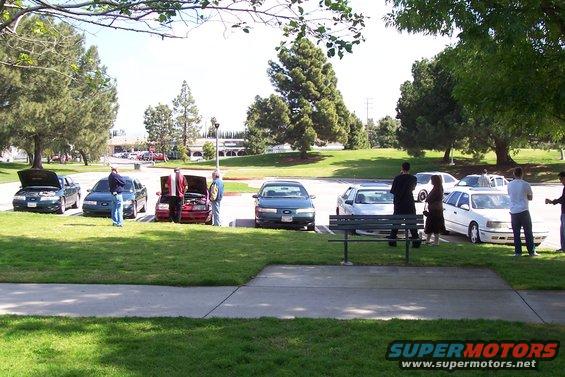 Several cars parked on grass and in a lot, with people standing around them. One car has its hood open