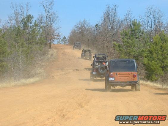 Four Jeeps driving up a dusty dirt road, one orange Jeep in foreground
