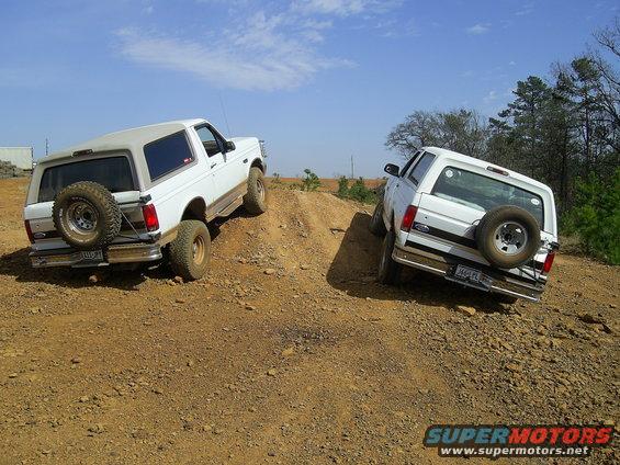 Two white Ford Broncos climbing a dirt hill, one behind the other