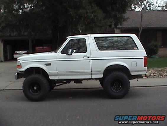 Lifted white SUV parked on a street, side view. It has large tires and appears to be a Ford Bronco from the early to mid-1990s