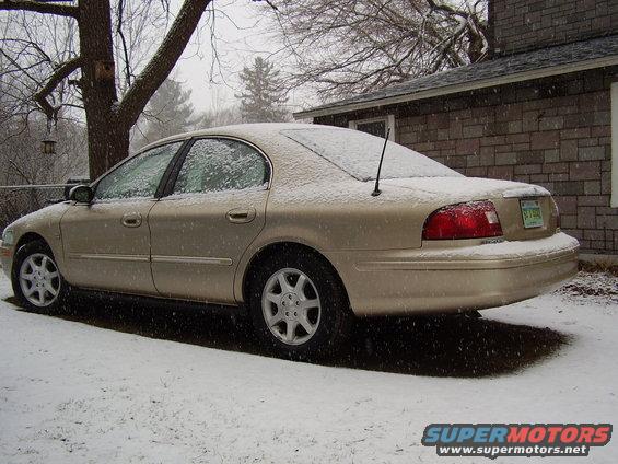 Gold sedan parked in snow, rear 3/4 view. Snow covers the car and ground. A brick house is in the background
