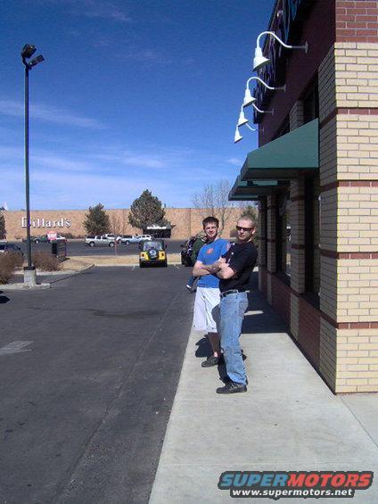 Two men stand on a sidewalk outside a brick building, with a yellow Jeep and other vehicles in the background