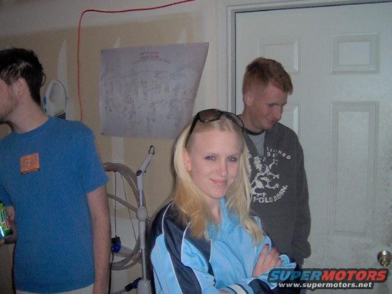 Three people stand in a garage with a poster on the wall and a white door